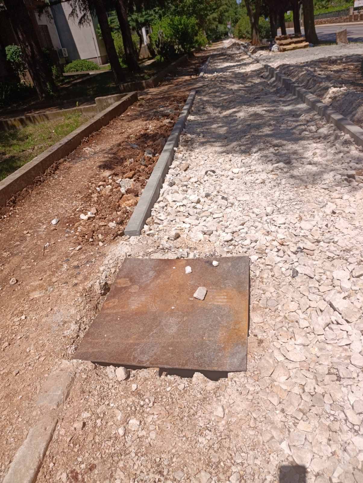 Razrovan nogostup ispred škole s otvorenom rupom pokrivenom metalnom pločom i nasipom šute